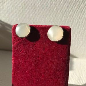 May include: A pair of white pearl earrings with silver settings. The earrings are displayed on a red velvet background.