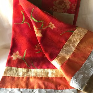 May include: A red and gold fabric table runner with floral patterns and embroidered details. The runner is made of multiple pieces of fabric sewn together, creating a patchwork effect.