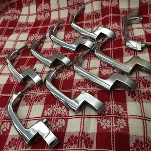 Op de afbeelding: Een set van acht verchroomde kastdeurgrepen. De grepen hebben de vorm van een lus met een recht gedeelte aan één kant. De grepen zijn in een verspringend patroon op een rood-wit geruite tafelkleed gerangschikt.
