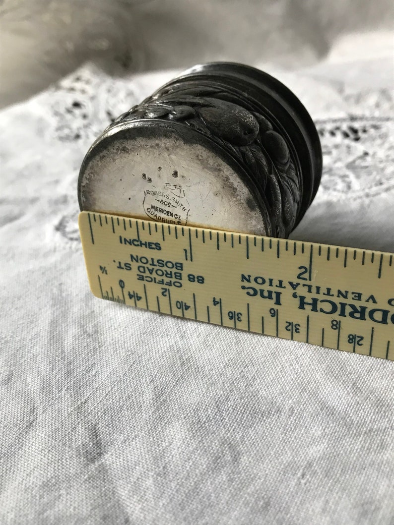 May include: A silver-plated, round, decorative box with an ornate design. The box is approximately 1 1/2 inches in diameter. The bottom of the box is marked with the text "Rogers Smith, Meriden, Ct. Quadruple."