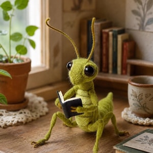Op de afbeelding: Een naaldgevilte groene sprinkhaanfiguur met grote zwarte ogen, lange voelsprieten en gesegmenteerde poten, die een klein open zwart boek met witte pagina's vasthoudt. De sprinkhaan is gemaakt van gestructureerde wolvezels.