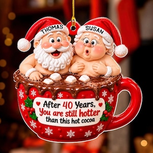 Op de afbeelding: Een hangende kerstversiering in de vorm van een rode mok gevuld met warme chocolademelk en marshmallows. De Kerstman en Kerstvrouw zitten in de mok. De mok bevat de tekst: After 40 Years, You are still hotter than this hot cocoa. Namen Thomas en Susan.