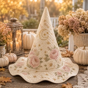 May include: A cream-colored quilted witch hat featuring embroidered pink and white floral patterns, gold crescent moon, and star motifs. The hat has a tall, pointed crown and a wide, wavy brim, constructed with detailed stitching throughout.