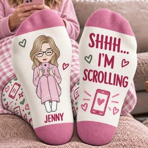 May include: A pair of white socks with pink cuffs and heels. The left sock features a cartoon illustration of a person with blonde hair and glasses holding a phone, with the name JENNY below. The right sock displays the text SHHH... I'M SCROLLING.