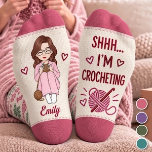 May include: A pair of cream-colored socks with dark pink cuffs and toes. The left sock features a cartoon illustration of a person with brown hair and glasses crocheting, with the name Emily below. The right sock displays the text SHHH... I'M CROCHETING with a yarn ball and hook graphic.