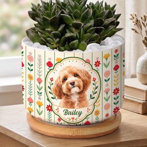May include: A cylindrical white planter featuring a vertical striped pattern with colorful floral illustrations and a central oval frame containing a portrait of a light brown dog. The name Bailey is printed below the dog. It sits on a wooden base.