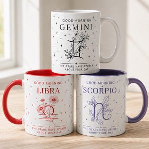 May include: Three white ceramic mugs featuring zodiac signs. The top mug says Gemini with floral art. The bottom left mug has a red handle and says Libra with floral art. The bottom right mug has a purple handle and says Scorpio with floral art.