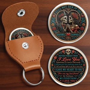 May include: A tan leather keychain pouch with a silver snap button, containing a round metal coin featuring two skeletons in formal attire and the text STILL NOT SICK OF YOU, DANIEL, and SOPHIA. A second matching coin displays the text I LOVE YOU.