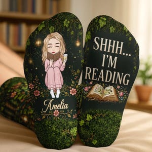 May include: A pair of dark green socks featuring a cartoon illustration of a person with blonde hair reading a book, the name Amelia, and the text SHHH... I'M READING above an open book graphic, all surrounded by vines, leaves, and small flowers.