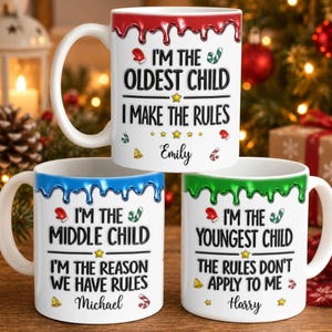 Könnte beinhalten: Drei weiße Keramiktassen mit farbigen Tropfenmustern am oberen Rand. Die obere Tasse hat rote Tropfen und den Text I'M THE OLDEST CHILD I MAKE THE RULES Emily. Unten links sind blaue Tropfen und der Text I'M THE MIDDLE CHILD I'M THE REASON WE HAVE RULES Michael. Unten rechts sind grüne Tropfen und der Text I'M THE YOUNGEST CHILD THE RULES DON'T APPLY TO ME Harry.