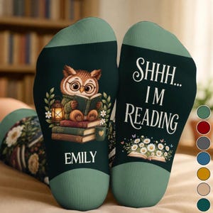 May include: A pair of dark green socks featuring a cartoon owl wearing glasses and reading a book atop a stack of books with a lantern. The text reads SHHH... I'M READING and EMILY. White daisies and butterflies are printed at the bottom.