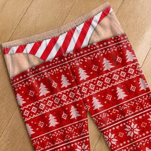 May include: Leggings featuring a red and white knit-style pattern with Christmas trees and snowflakes. The waistband area displays a printed graphic of skin-toned fabric and a red and white diagonal striped thong-style undergarment design.