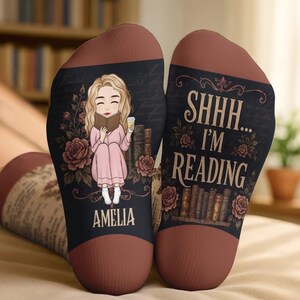 May include: A pair of black socks with a brown toe and heel. The left sock features an illustration of a blonde person reading a book with the name AMELIA printed below. The right sock displays the text SHHH... I'M READING above a stack of books.