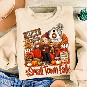 May include: A beige sweatshirt featuring a graphic of a cartoon character in a brown sweater standing by a red truck. The design includes a wooden sign reading 47567 PETERSBURG, a barn, pumpkins, and the text Small Town Fall and PUMPKINS HAYRIDES APPLE CIDER.