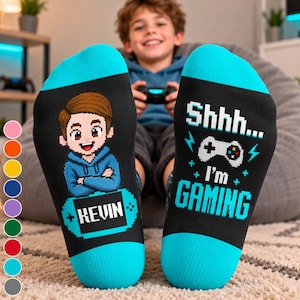 Puede incluir: Un par de calcetines negros con puños azul brillante. El calcetín izquierdo muestra un personaje pixelado con sudadera azul sobre una consola portátil con el nombre KEVIN. El derecho muestra el texto Shhhh... I'm GAMING y un icono de mando.