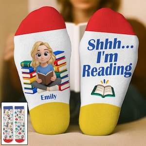 May include: A pair of white socks with red toe and yellow heel sections. The left sock features a cartoon illustration of a blonde person reading a book next to a stack of books with the name Emily. The right sock displays the text Shhh... I'm Reading.