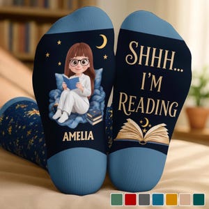 May include: A pair of dark blue socks featuring a cartoon character with brown hair and glasses reading a book while sitting on a blue cushion. The text reads AMELIA and SHHH... I'M READING, accompanied by an open book graphic and a crescent moon.