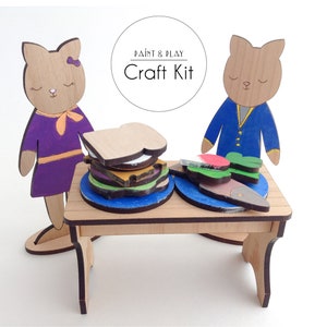 May include: A wooden craft kit featuring two cat figures, a table, and pieces to create a sandwich and a plate of food. The kit is unpainted and ready to be decorated. "Paint & Play Craft Kit" is written on a circle above the figures.