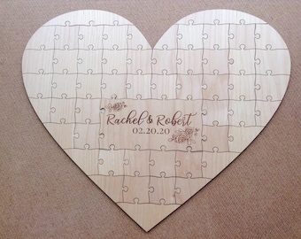 May include: A wooden heart-shaped puzzle with interlocking pieces. The puzzle is engraved with the names "Rachel & Robert" and the date "02.20.20" in a decorative font. The puzzle is made of light-colored wood.