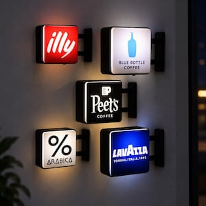 Puede incluir: Cinco letreros cuadrados iluminados montados en la pared con logotipos de marcas de café. Los letreros muestran los nombres illy, Blue Bottle Coffee, Peet's Coffee, % Arabica y Lavazza Torino, Italia, 1895. Cada letrero tiene un soporte negro.