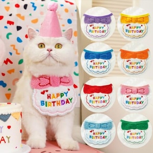 以下が含まれることがあります： ピンクの誕生日の帽子と「Happy Birthday」の文字が入ったよだれかけをつけた白い猫。画像には、リボンとテキストが同じカラフルなよだれかけもいくつか表示されています。小さなケーキが見えます。