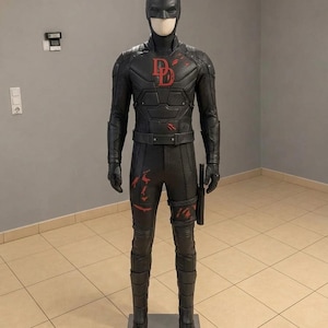 Puede incluir: Un disfraz completo de Daredevil en color negro sobre un maniquí. El traje presenta un acabado texturizado, un logotipo DD rojo en el pecho y detalles rojos en el torso y las piernas. Incluye una máscara con cuernos, un cinturón y una funda.