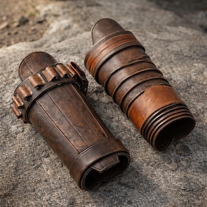 May include: A pair of dark brown leather arm guards featuring a textured finish. One guard includes a series of small loops along the top edge, while the other displays a layered, wrapped design with multiple horizontal bands and a smooth surface.