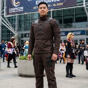 May include: A person in a brown costume stands in front of a convention center. The costume includes a high-collared top and matching pants with visible stitching. The banner reads "COSPICON 2024 - MAIN EXHIBIT HALL ENTRANCE OCT 10-18, 2024."