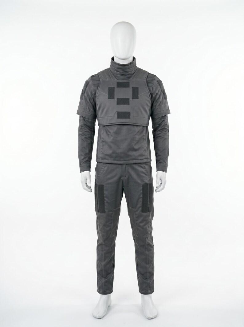 Op de afbeelding: Een grijs tactisch uniform gepresenteerd op een mannequin. De outfit omvat een shirt met lange mouwen en een hoge kraag, een vest met vierkante patches en een bijpassende broek. Het uniform is ontworpen voor bescherming en duurzaamheid.
