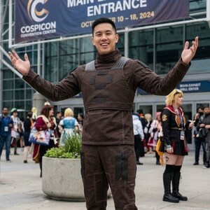 May include: A person in a brown cosplay outfit poses in front of a banner for COSPICON 2024. The outfit includes a high-collared top, pants, and boots. The banner reads "COSPICON 2024 - MAIN EXHIBIT HALL ENTRANCE OCT 10-18, 2024."