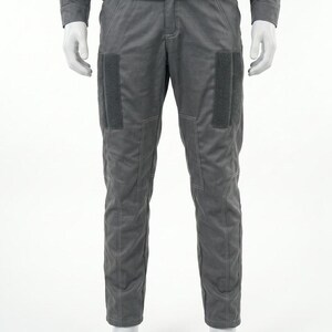 Op de afbeelding: Een grijze jumpsuit gepresenteerd op een mannequin. De outfit bestaat uit een top met lange mouwen en een broek met grote rechthoekige patches op de dijen. De broek heeft een getailleerd ontwerp met zichtbare stikseldetails.