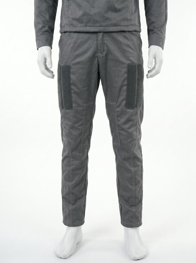 Op de afbeelding: Een grijze jumpsuit gepresenteerd op een mannequin. De outfit bestaat uit een top met lange mouwen en een broek met grote rechthoekige patches op de dijen. De broek heeft een getailleerd ontwerp met zichtbare stikseldetails.