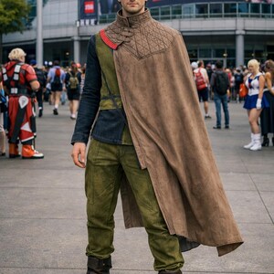 Puede incluir: Un cosplay con capa marr&oacute;n, pantalones verde oliva y botas marrones oscuras. El atuendo incluye un cuello acolchado y un acento rojo. El fondo muestra un centro de convenciones con la inscripci&oacute;n "WELCOME TO ANIME EXPO!"