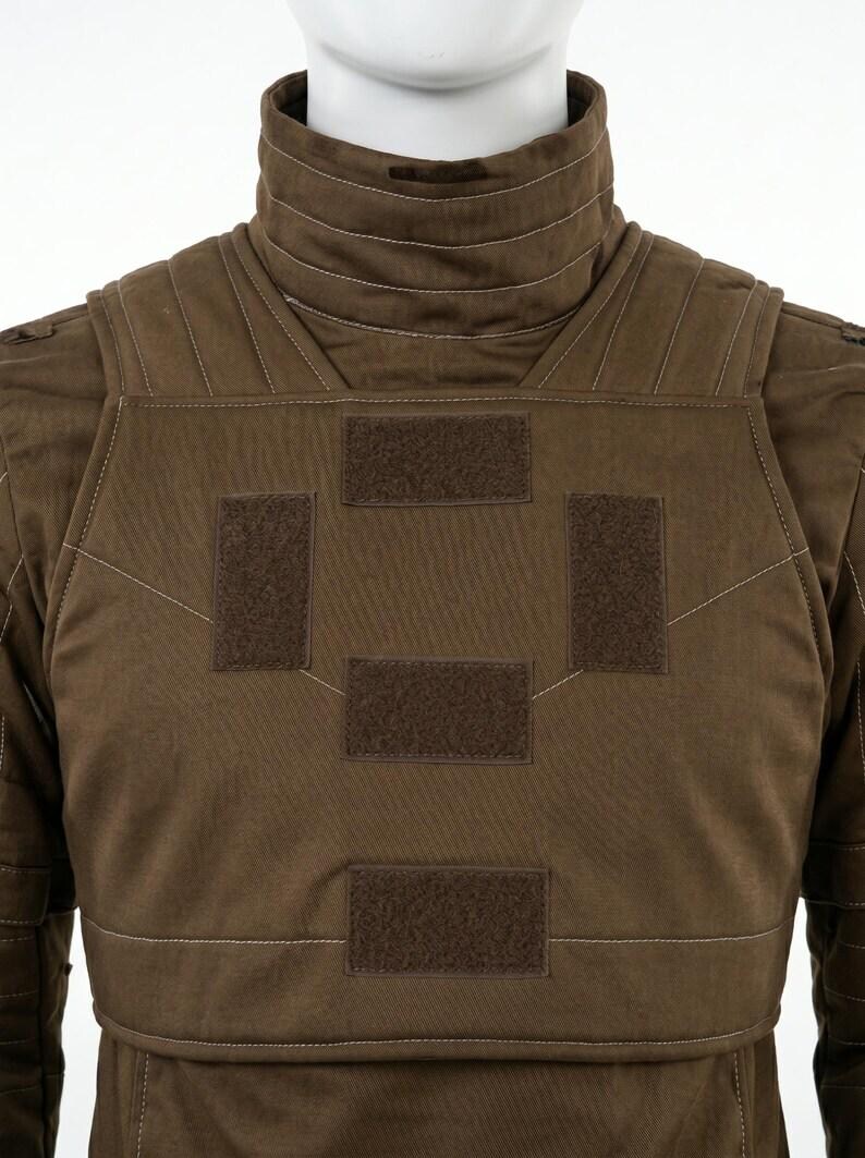 K&ouml;nnte beinhalten: Eine braune taktische Weste mit hohem Kragen und horizontalen N&auml;hten, strukturierten Schulterpartien und vier rechteckigen Klettverschluss-Patches auf der Brust. Das Kleidungsst&uuml;ck ist mit sichtbaren N&auml;hten verarbeitet.