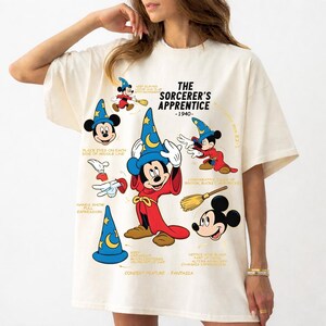 Può includere: Una t-shirt color crema con una stampa grafica di Topolino nei panni dell'apprendista stregone del 1940. Il design include diverse illustrazioni del personaggio, un cappello da mago, una scopa e testo didascalico che descrive le caratteristiche.