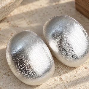 May include: A pair of silver-toned earrings with a smooth, rounded, oval shape and a brushed, textured metallic finish. The earrings feature a convex surface that reflects light, creating highlights across the curved, polished metal exterior.