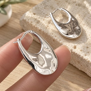 May include: A pair of silver hoop earrings with a polished, irregular, and wavy surface texture. Each earring features a teardrop-shaped design with a hinged closure mechanism. The metal is stamped with the hallmark 925, indicating sterling silver.