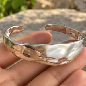 May include: A polished silver cuff bracelet with an irregular, organic, and wavy surface texture. The open-ended band features a stamped 925 hallmark on the interior, indicating sterling silver composition, with a reflective, high-shine finish.