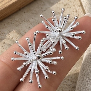May include: A pair of silver-toned earrings featuring a starburst design. Each earring consists of multiple thin, radiating metal rods that extend from a central point, with each rod ending in a small, smooth spherical bead, creating a sunburst effect.