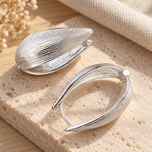 May include: A pair of silver-toned hoop earrings featuring a textured, leaf-like surface with fine vertical striations. The earrings have a hinged closure mechanism and an elongated, oval shape that tapers to a point at the bottom end.