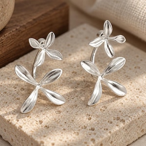 May include: A pair of silver-toned earrings featuring a double-flower design. Each earring consists of two stacked, four-petaled floral shapes connected by a curved metal stem, creating a layered, dimensional appearance with a polished metallic finish.