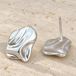May include: A pair of silver-toned stud earrings with an irregular, organic, and rippled surface texture. One earring is positioned face-up, while the other is turned to show the metal post backing used for securing the jewelry to the ear.