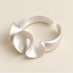 May include: A silver-toned ring featuring an open band design with three distinct, curved, petal-like shapes extending from the front. The metal surface has a matte, brushed finish, creating a soft, non-reflective appearance across the entire piece.