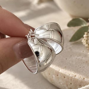 May include: A pair of polished silver hoop earrings with a wide, curved, and reflective surface. The earrings feature a hinged closure mechanism at the top, showcasing a smooth, metallic finish that catches the light, creating a bright appearance.