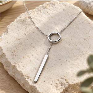 May include: A silver-toned lariat necklace featuring a delicate chain that passes through a circular ring, ending in a smooth, rectangular bar pendant. The metal has a polished, reflective finish that catches the light, creating a clean, linear aesthetic.