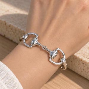 May include: A polished silver-tone metal bracelet featuring a central horsebit-inspired design. The chain consists of two symmetrical, curved metal loops connected by a horizontal bar, creating a continuous, sleek, and reflective link structure.