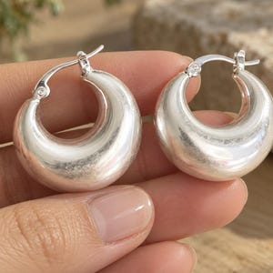 May include: A pair of polished silver-tone hoop earrings featuring a thick, rounded, crescent-shaped design. The earrings have a smooth, reflective surface and a hinged post closure mechanism at the top, creating a continuous circular silhouette.