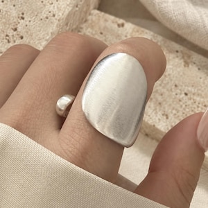 May include: A silver-toned ring featuring a large, flat, organic-shaped circular plate with a brushed matte finish. The ring has an open-band design, showcasing a smooth, rounded metal segment on the side of the finger, creating a bold look.