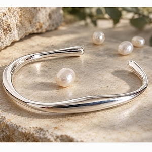 May include: A polished silver cuff bracelet with a smooth, organic, and slightly irregular curved shape. The bracelet is accompanied by three small, round, white freshwater pearls placed near the open ends of the metal band.