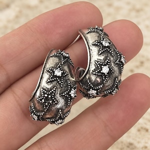 May include: A pair of silver-toned hoop earrings with a textured, oxidized finish. Each hoop features multiple raised star shapes, with a small, clear, round-cut stone set in the center of each star, creating a celestial-inspired design.